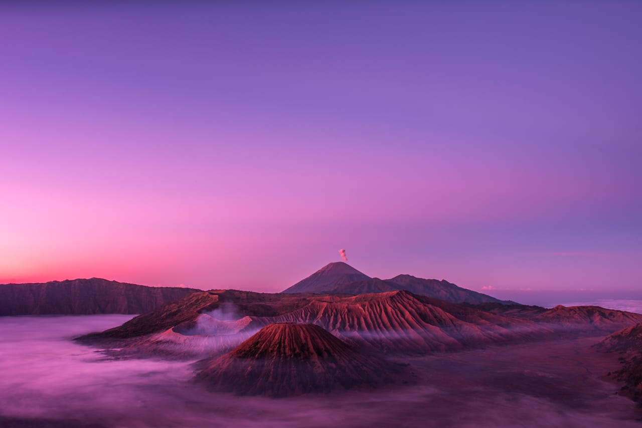 Sebuah kawah gunung berapi dalam cahaya pertama pagi, awan dan langit berwarna magenta dan ungu.