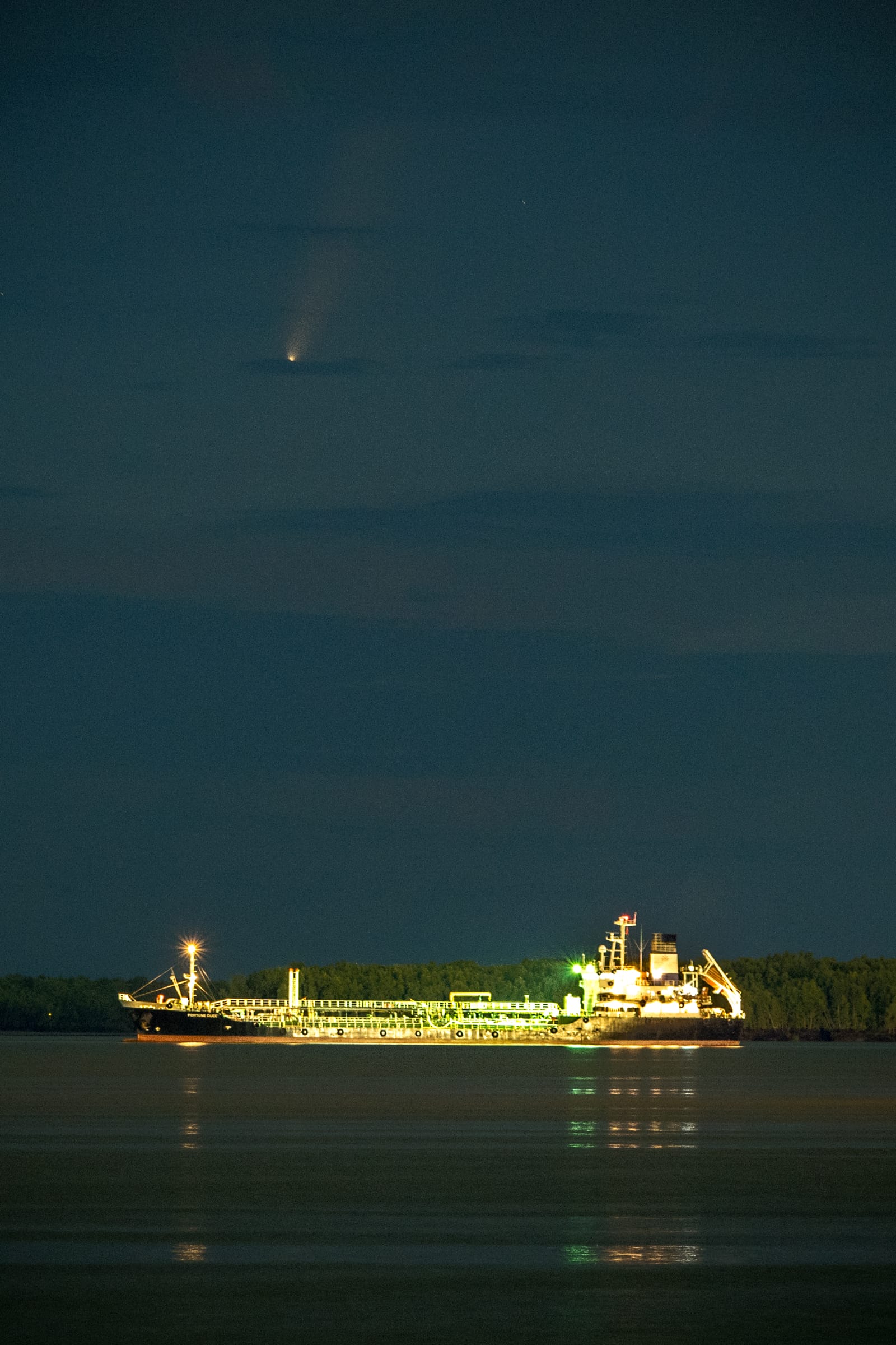 Portrait frame: a lit cargo ship on the night sea, a comet trailing its tail above it.