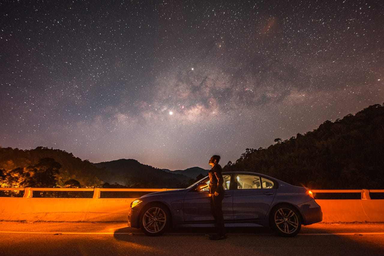 夜里的水坝公路上，Kevin 站在车旁仰望银河，钠灯把护栏照成橙色。