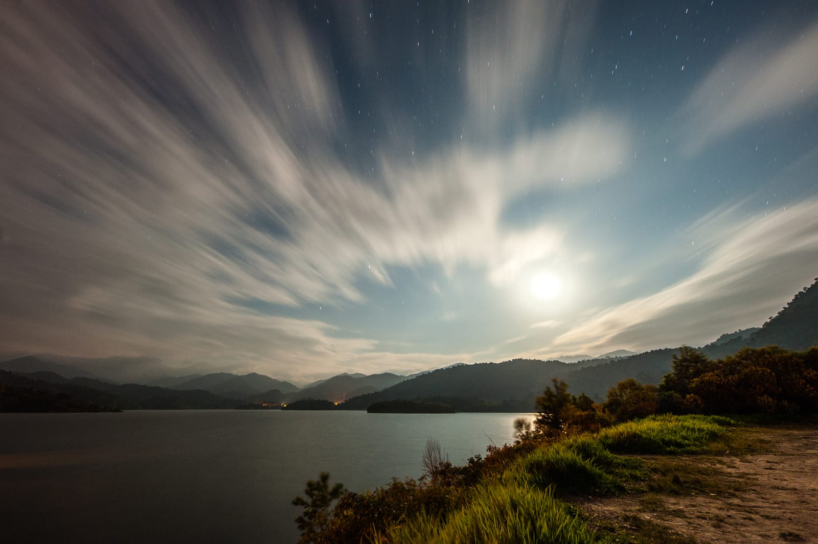 The moon above a lake; a long exposure draws the moving clouds into streaks of light.