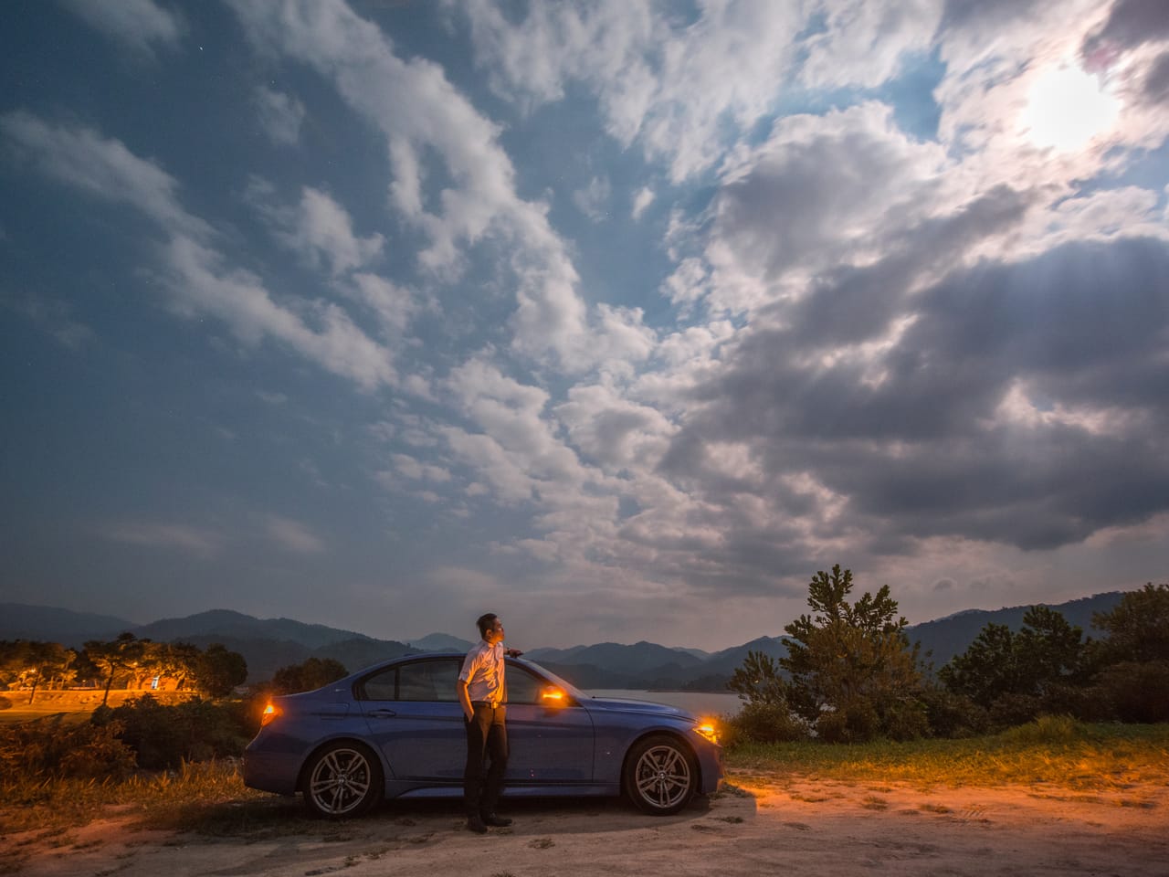 Moonlit clouds fill the sky; Kevin, in a white shirt, stands beside his car on a lit hillside.