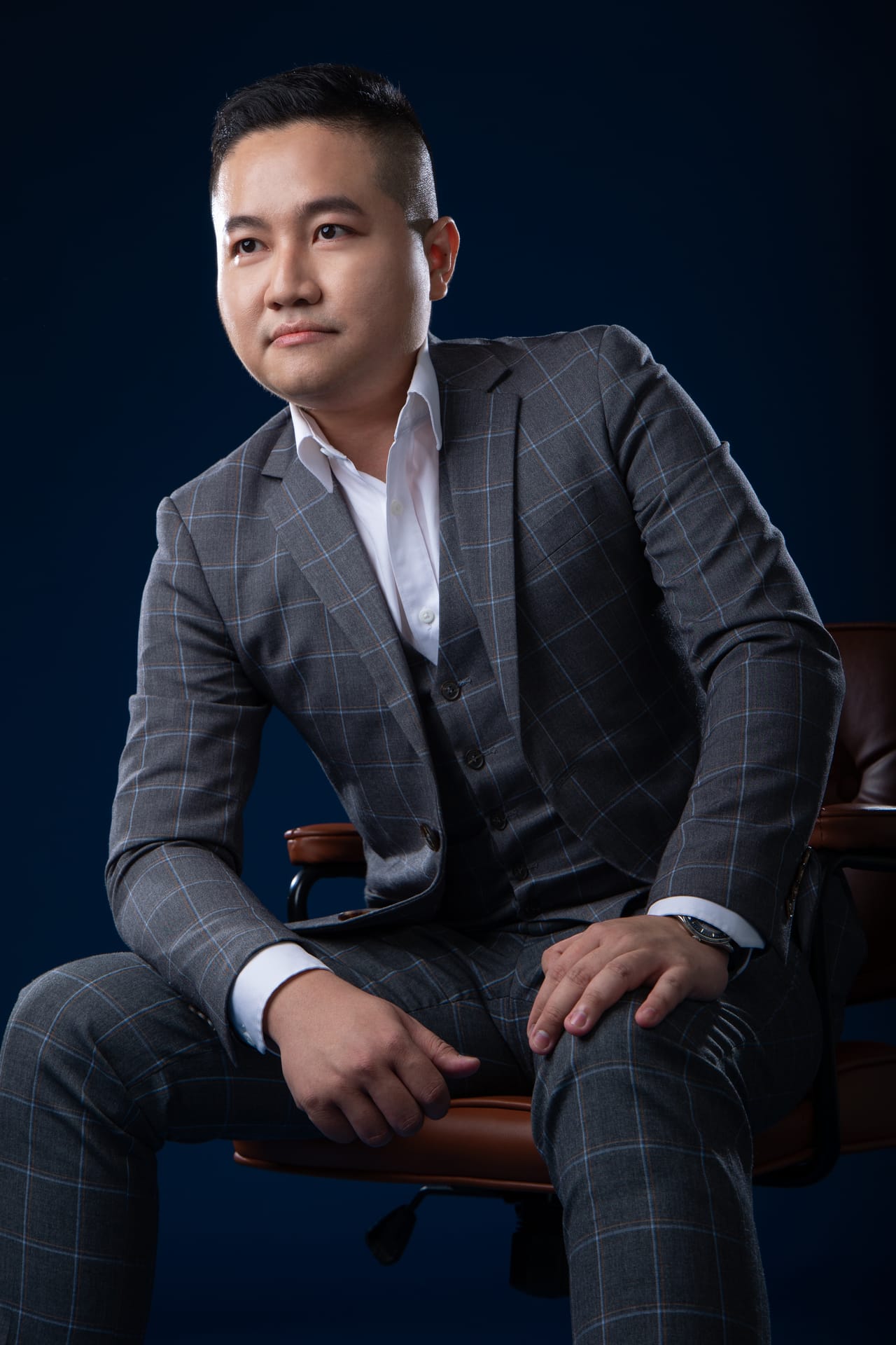 深蓝背景前，穿灰色格纹西装的 Kevin 坐着，身体前倾，手肘搭在膝上，看着镜头。