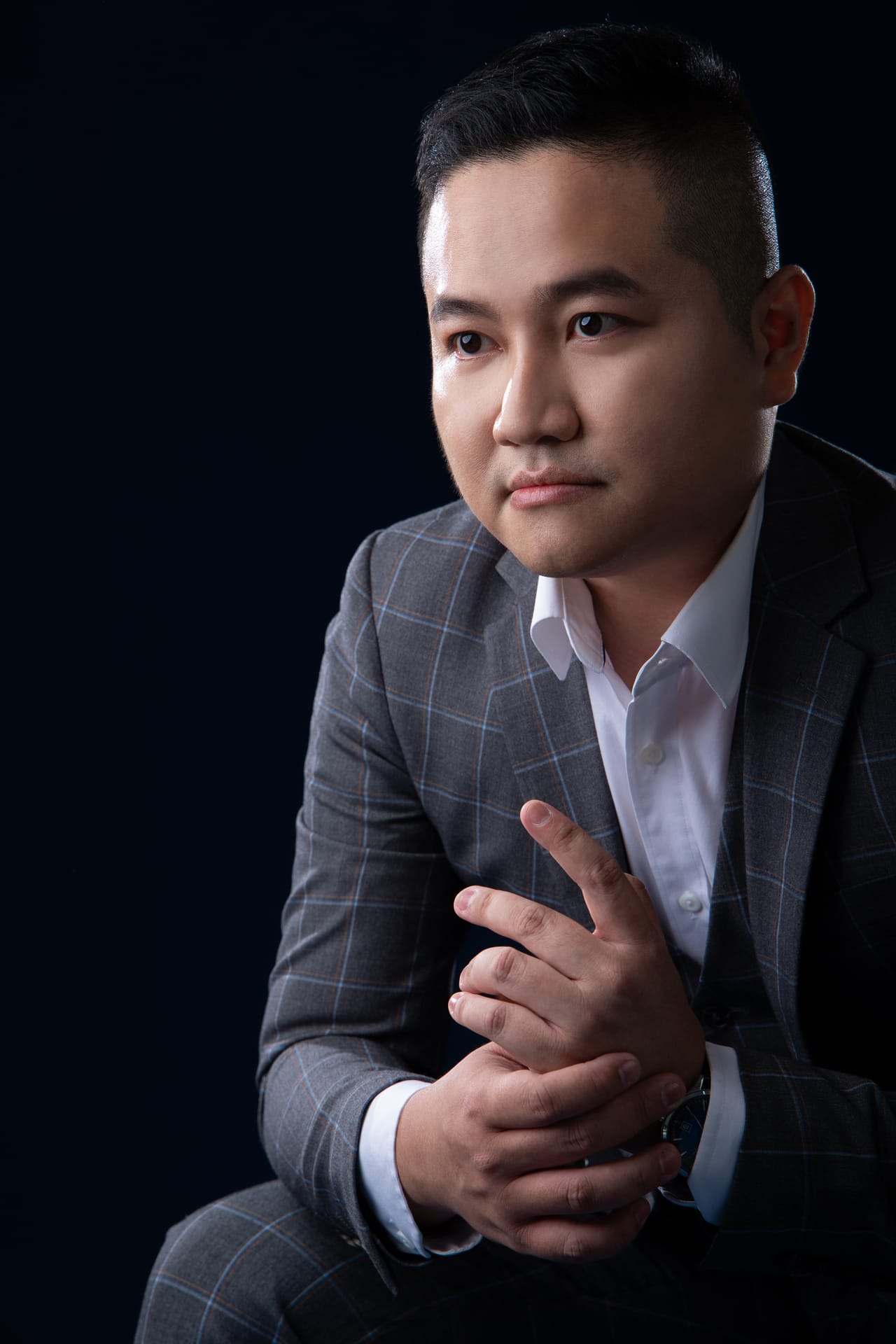 Berlatarkan biru tua, Kevin duduk dengan tangan digenggam perlahan, memandang ke kamera dengan penuh perhatian.