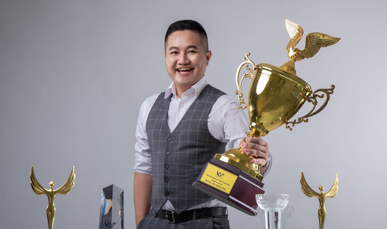 浅灰背景前，穿灰色格纹背心的 Kevin 笑着举起一座 FLP Realty 的大奖杯，铭牌写着 TOP ADVISOR OF THE YEAR 2019，两旁立着金色的奖座。