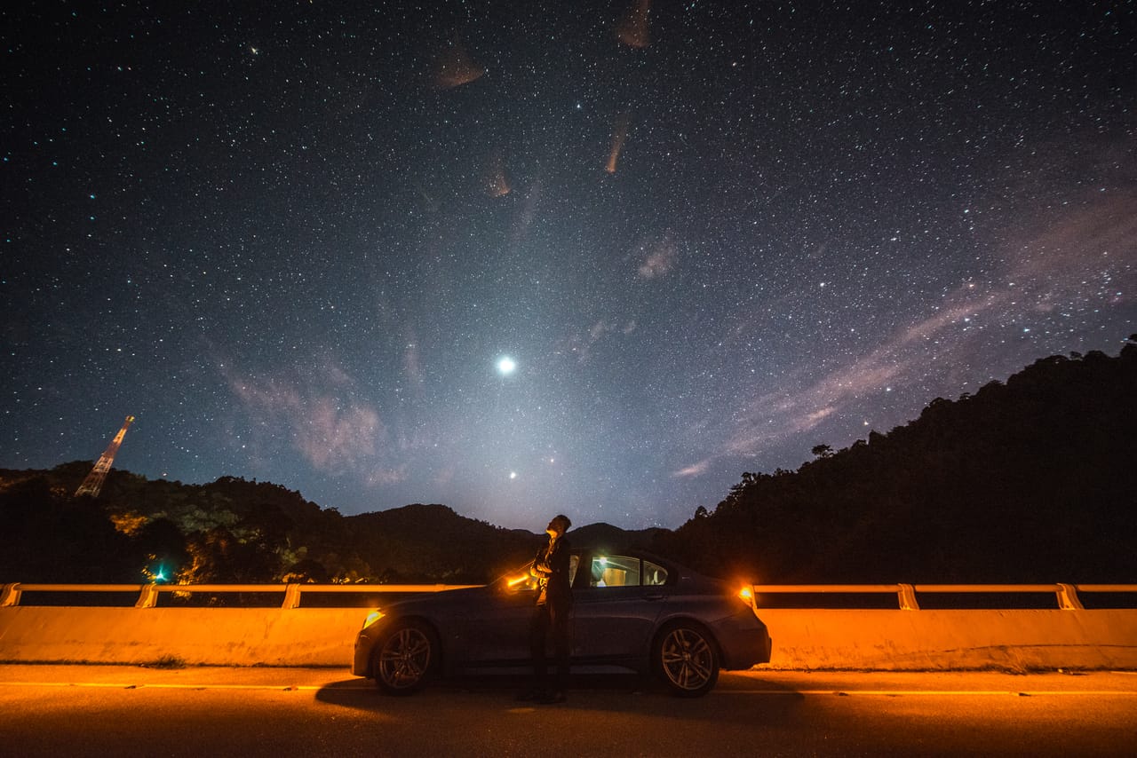 A dam road at night: sodium lamps light the guardrail orange as Kevin stands by his car under a sky full of stars.