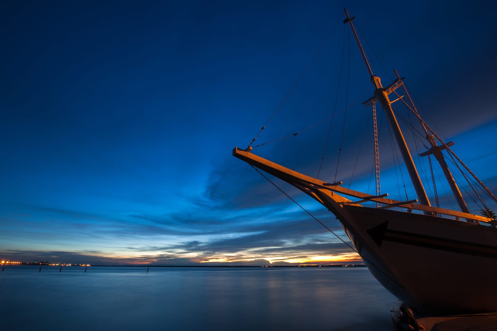 Blue hour: the bow of a wooden sailing ship reaches into the frame; sunset and lights on the far horizon.