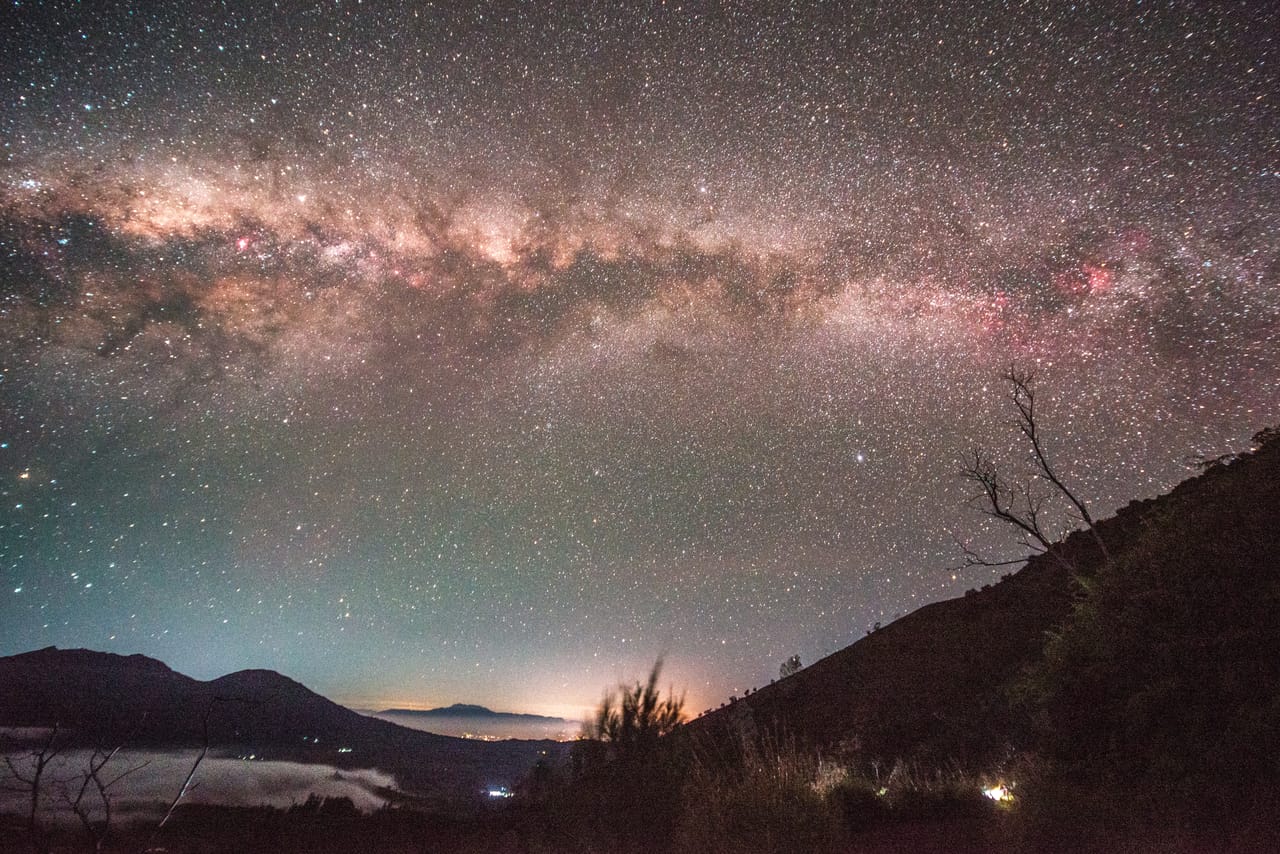 The Milky Way arches across the night over a sea of clouds and the outline of a volcano; a dead tree stands on the slope.