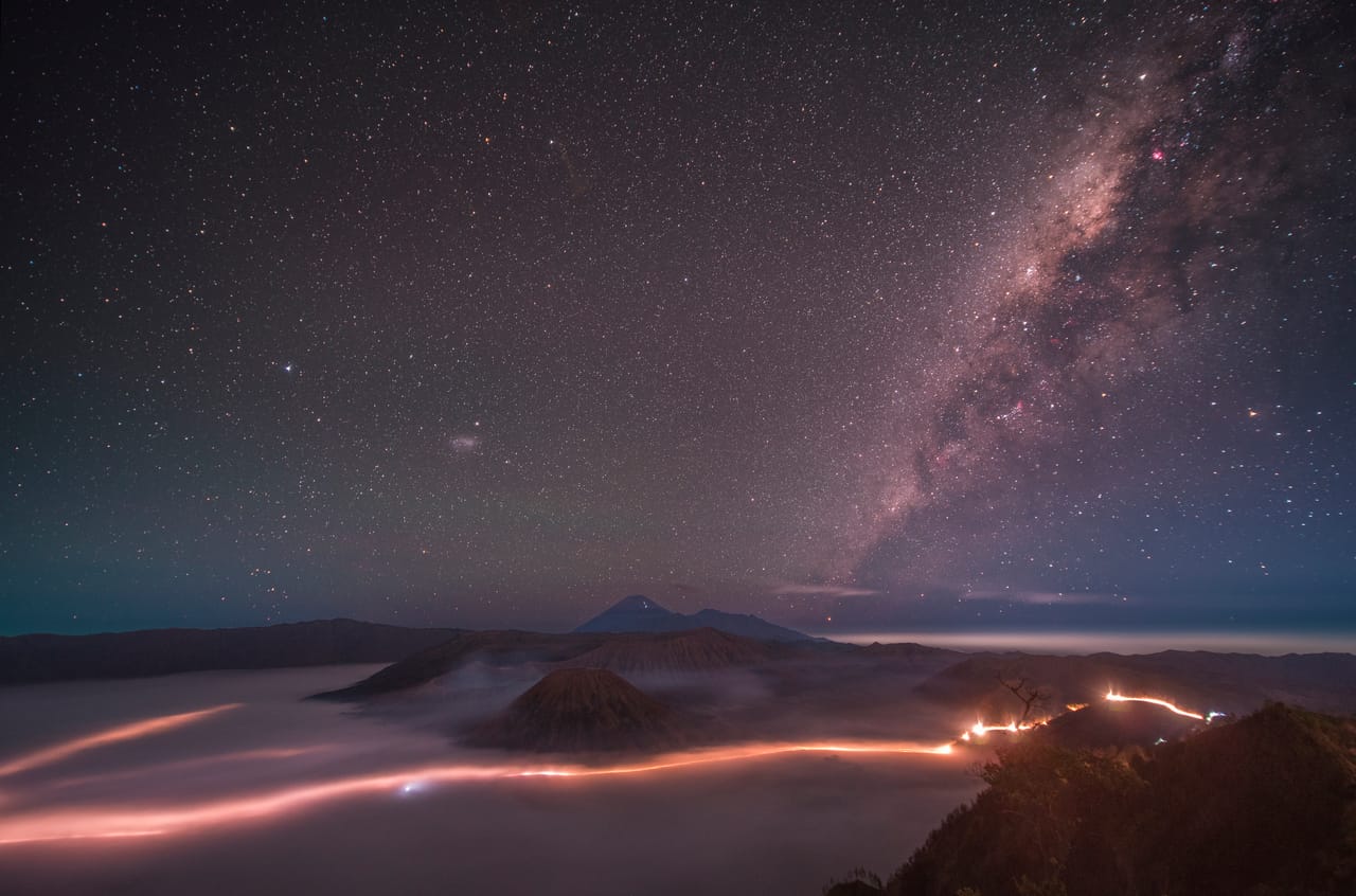 A sea of clouds fills the valley; headlights draw a line of light beneath it; the Milky Way slants across the upper right.