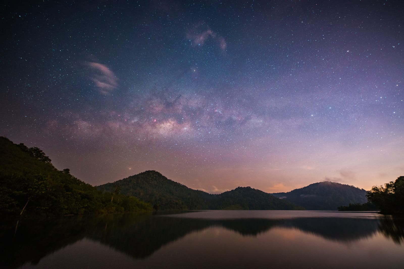The Milky Way crosses the sky above a lake, the hills and stars reflected in the water.
