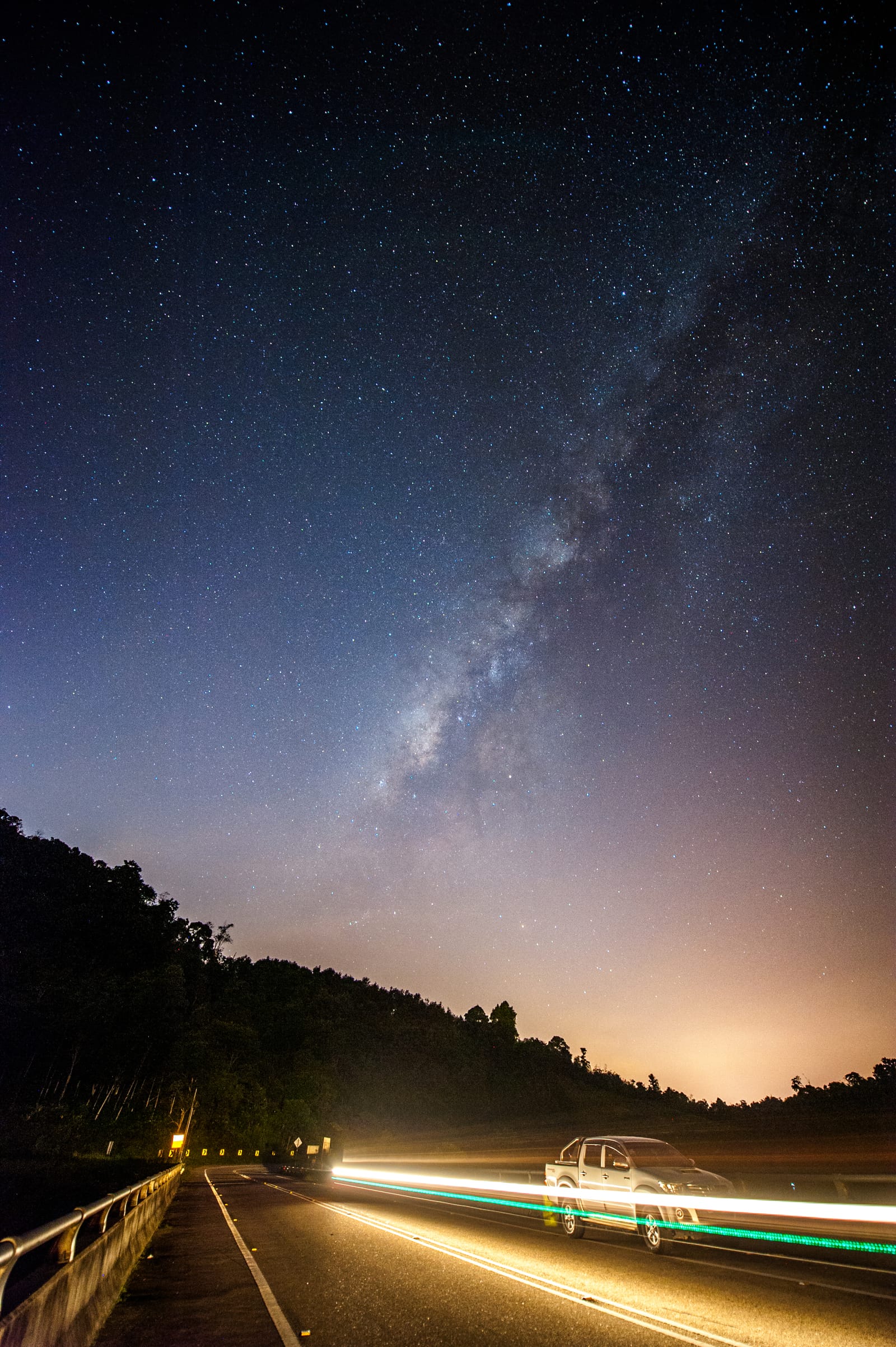 Portrait frame: the Milky Way rises over a mountain road as a passing car leaves a long trail of light.