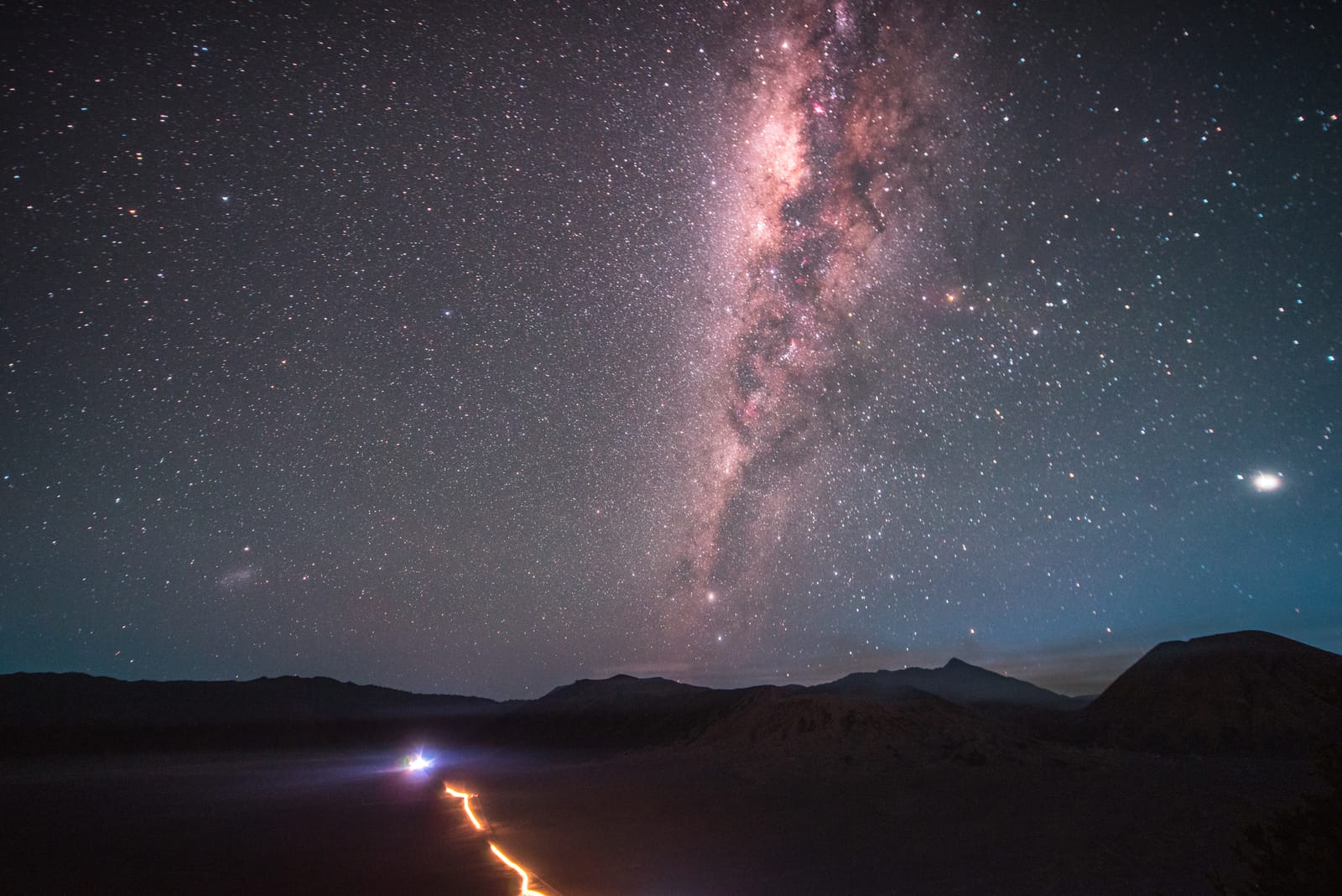 The Milky Way rises straight up while an orange trail of headlights winds up the valley.