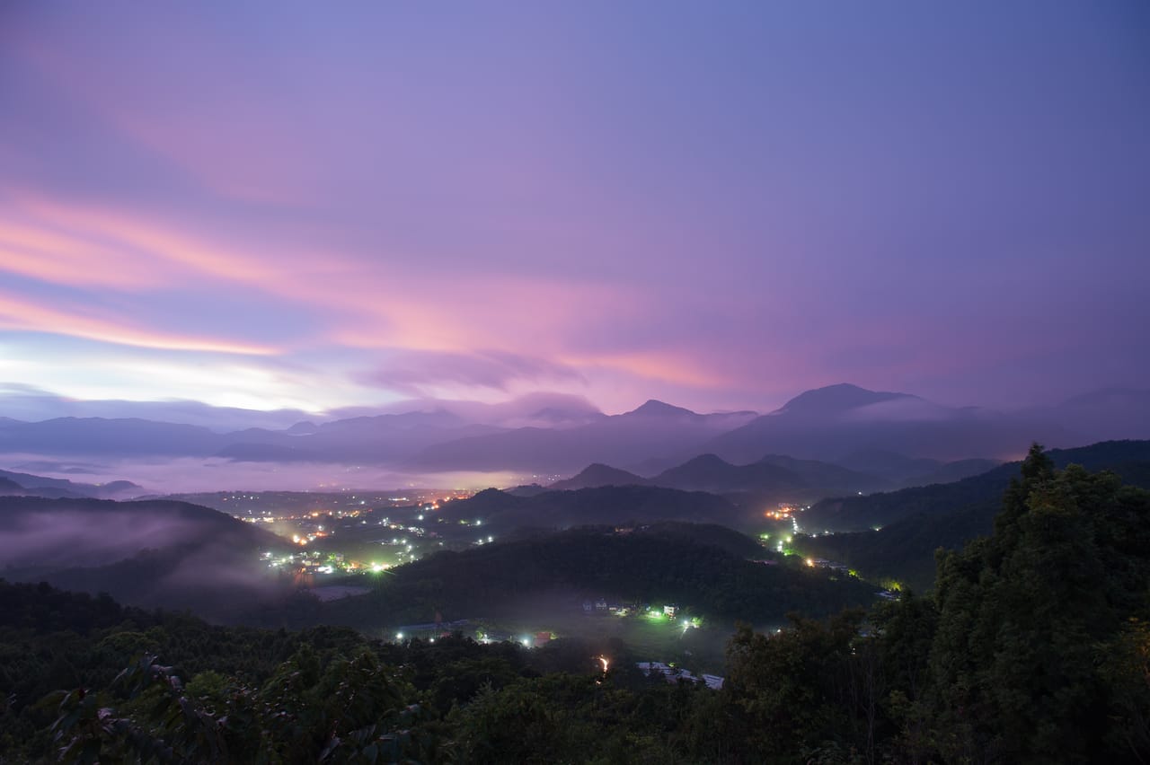 A valley at dusk: a small town’s lights glow through the mist under a pink and violet sky.